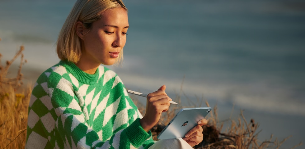 A person on the beach holding their Apple Pencil Pro in one hand and a blue iPad mini in the other
